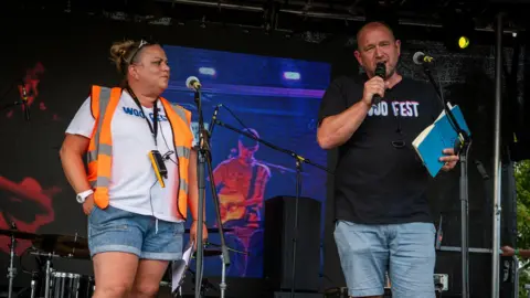 Ria and Luke Simons pictured on stage. Ria Simons is wearing an orange high viz jacket with a white top and denim shorts. Luke Simons has a black t-shirt with the words "Woo Fest" written on the front and is wearing blue shorts.
