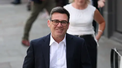 Reuters Andy Burnham smiling while walking in a black suit and white shirt.