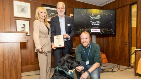 Our Bury St Edmunds Two men and a woman posing with black dog while holding an award