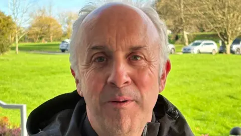 A man with grey hair in a dark jacket standing in front of some grass with parked cars in the distance.