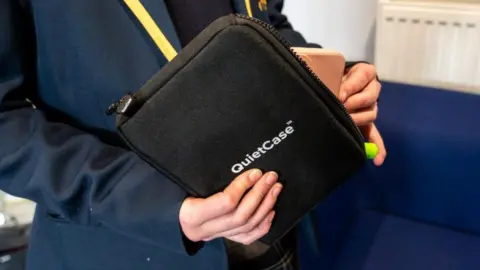 A close-up of a girl in a navy school blazer holding a black pouch with a zip on the side. "Quiet Case" is written in white lettering on the side. 