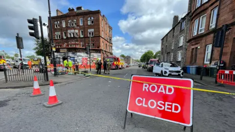 A road closed sign and police tape block off a city street beside a junction lined with red sandstone tenement buildings. Emergency service workers, including firefighters and police officers, stand near fire engines and cordoned-off pavement areas under a partly cloudy sky.