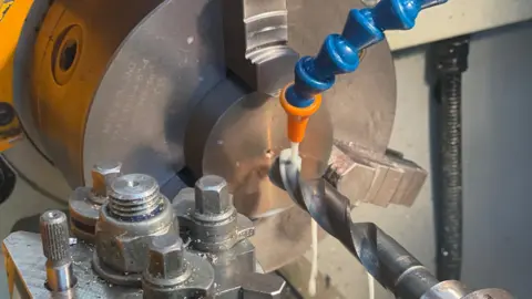 James Goh A close-up shot inside a workshop shows a piece of metal being worked on using machinery. The metal component is clamped in a rotating chuck, while a large drill bit presses into its centre. Liquid coolant is flowing over the area to reduce heat and friction during the process. The image highlights the precision engineering involved in manufacturing the fidget spinner.
