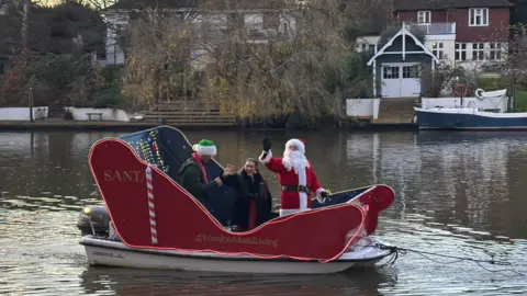 Hannah Karpel/BBC A sleigh mounted on a small motorboat carries people dressed as Father Christmas and two others along the River Thames, with houses and moored boats visible on the far bank.