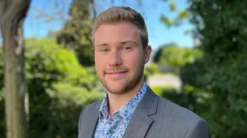 Simon Dedman/BBC Joe Holmes stands outside on a sunny day in front of several bushes and smiles at the camera. He has short blond hair and wears a grey suit jacket with a blue floral shirt underneath.