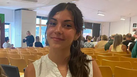 Sofia sits in a school hall. She has long, straight dark-brown hair which is tied up in a ponytail and is wearing a white sleeveless top.