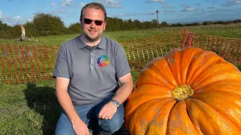 Ben Rayner Ben Rayner sits on the grass next to his huge pumpkin. He leans against it. He is wearing a gray shirt and sunglasses.