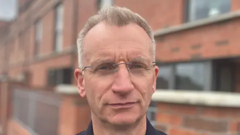 Kingston has short grey hair and eyebrows, he is wearing clear and metal rounded glasses. He is pictured in a close-up shot and is wearing a black jacket.