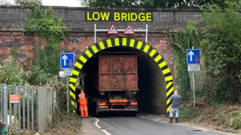 Martin Heath/BBC Back of a red lorry is visible inside a bridge under the sign "Low Bridge". A man in orange overalls and another man in a grey shirt are looking at the lorry.