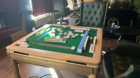 A gambling table with chips on it, there are chairs around it and windows with the curtains drawn in the background.