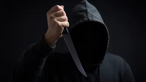 A man is wearing a black hooded top with his face hidden and is holding a knife