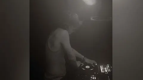 Shaggy FX A black and white image of a young DJ playing a record. The man has short, fair hair and is wearing headphones on his head. He has a vest on.