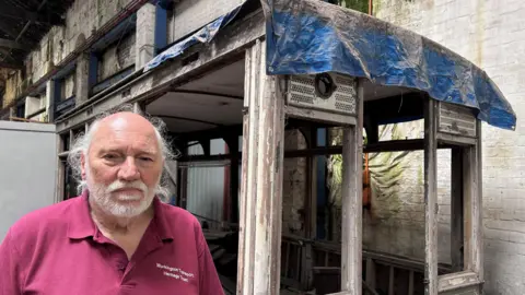Philip Tuer stands in front of the shell of the old tram, looking at the camera. He has a white beard and moustache, and thinning, longish hair, pulled back. He is wearing a red polo shirt. The wooden shell of the dishevelled tram carriage stands in a white-bricked warehouse. There is blue tarpaulin placed over the roof, and wooden panels piled up inside.