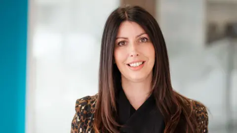 Hudgell Solicitors Solicitor Hayley Collinson. She has long brown hair and brown eyes. She is wearing a leopard print blazer over a black blouse. She is smiling at the camera and the background is blurred.
