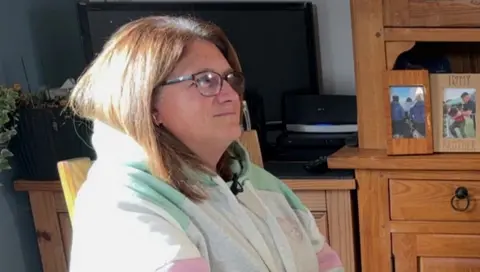 Nicole Bell-Percy wearing a grey, hooded jumper and glasses, sits in a chair