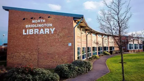 A crescent-shaped, brick-built building in a landscaped garden. There is a sign on the side of the building that reads: North Bridlington Library.