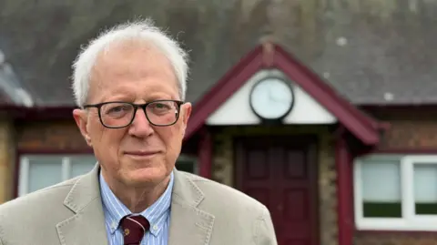 A elderly man stood in from of a maroon painted doorway that has an analogue clock above it. The man has grey thinning hair and glasses. He is wearing a cream coloured blazer with a striped canvas shirt and a maroon tie.