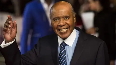 Getty Images Paul Stephenson in a blue shirt, navy and pale blue striped tie and a black jacket, smiling at the camera and holding done hand up in a wave. 