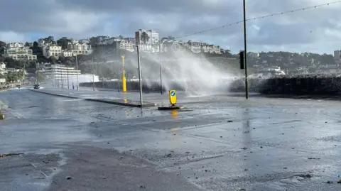 A picture of a road which has been covered in flood water. Large waves can be seen over the top of the sea wall.