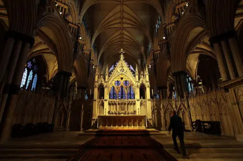 Getty Images The interior of a large gothic cathedral in semi-darkness, looking up towards vaulted ceilings. The view is dominated by the high altar, with highly decorative stonework lit up brightly amid the surrounding gloom. In the foreground, the figure of a man, in shadow, can be seen walking up steps.