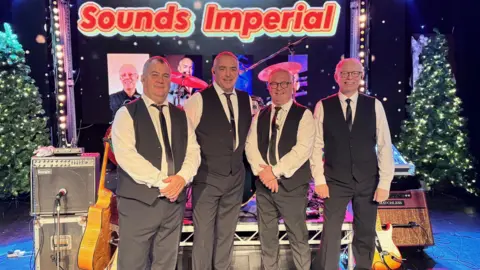 Supplied Four men wearing black suits and white shirts standing in front of a small stage and a sign saying Sound Imperial