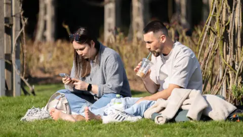 PA Media A woman and a man sit in the sunshine on some grass. Both have their trainers off. The man is drinking and iced-coffee and the woman is reading her phone.