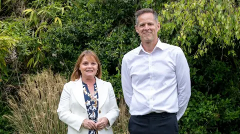 A woman with shoulder length brown hair, wearing a blue and white dress and a white jacket, is standing looking at the camera. Next to her is a man with grey hair and wearing a white shirt and black trousers. Both are smiling. Behind them are green bushes and trees