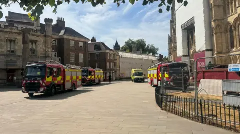 BBC/Emily Johnson Three red fire engines and a yellow ambulance are parked beside historic stone buildings, one covered in scaffolding and white sheeting, with a “Danger Keep Out” sign on a fence in the foreground under a bright, clear sky.