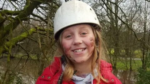 Family photo A younger Menna smiles at the camera. She has long blonde hair, wears a white helmet and red coat, and has mud on her cheeks. A river and trees can be seen in the background. 