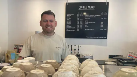 Bradley Buechel wears a grey t-shirt and looks into the camera from behind his professional coffee machine. A price list is behind him on the wall and there are coffee cups lined up on the top of the machine.