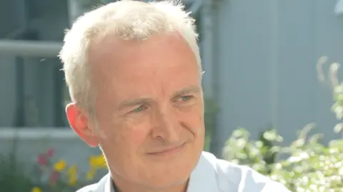 Jamie Niblock/BBC Dr Ewen Cameron smiles while looking away from the camera. He sits outside where bushes and flowers can be seen behind him. He has short grey hair and wears a white shirt. 