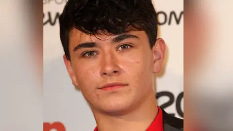 Close up of actor William Rush with dark brown hair and hazel eyes wearing a red top and black jacket at an awards ceremony.