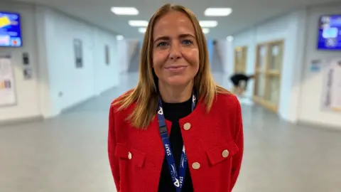 BBC / Christian Johnson stands in a bright, white atrium of a head teacher school hall. Her shoulder has golden hair, wears a shiny red suit jacket and smiling on the camera.