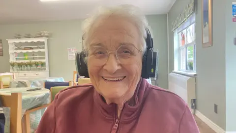 An older woman, with short grey hair, wearing headphones and a dark pink zip-up jumper.