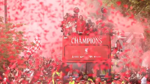 EPA Image shows Liverpool FC's victory parade bus moving through Liverpool. Players stand on the open top deck as thousands of fans surround the vehicle. Confetti and smoke can be seen hanging in the air. 