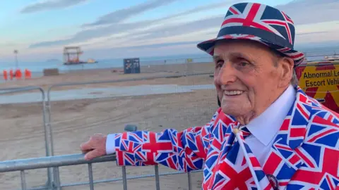 An elderly man pictured in front of a beach wearing a suit covered in Union flags and wearing a hat with the same pattern on it.