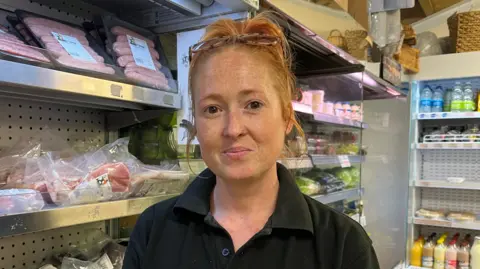 Laura looking into the camera and smiling slightly. She has tied up red hair with glasses on her head and is wearing a black short-sleeved polo shirt, standing in front of a row of stainless steel, cold shelves. On the shelves are various packets of meat; sausages on the top rack and bacon in the middle. Behind her there are also shelves holding fresh vegetables and fruit juice.