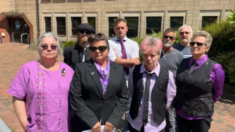 The family of Bernadette stood outside of the court all wearing purple. There are eight people stood together, with some wearing sun glasses.