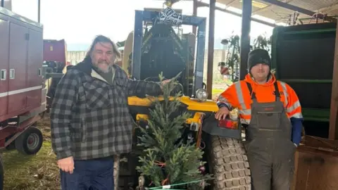 Julia Hubbard Two men stand either side of a tractor, smiling at the camera, with a Christmas tree in the middle of them. The man on the left has shoulder length dark hair and a grey beard. He is wearing a lumberjack style shirt. The man on the right has a dark coloured beanie hat and an orange high visability hoodie, with dungarees.