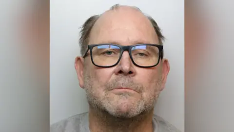 A mugshot of Martin Chubb, who is wearing a grey T-shirt. He is unshaven and balding. He has dark glasses.