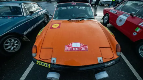 Susie Mackenzie/SDM Photography Three classic cars parked in a car park. They are dark green, bright orange and red