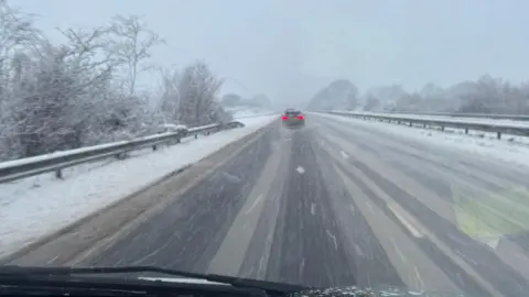 Spencer Clark Snow falls on the A30 in Cornwall near the Jamaica Inn pub. The road has a light scattering of snow on it with tracks from where vehicles have driven clear to see. A car is in the distance with its brake lights on.