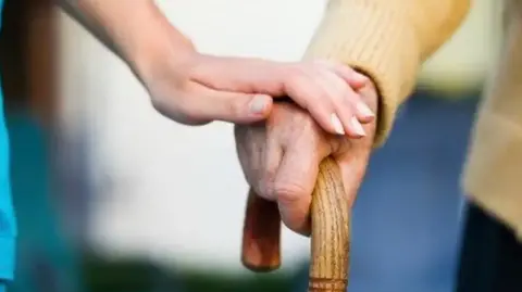 A woman places her hand on a an older man's hand as he holds a wooden walking stick.