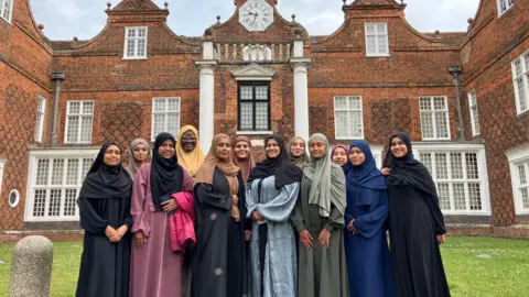 Jon Wright/BBC 12 women wearing traditional Muslim dress with headscarves stand in front of Christchurch mansion in Ipswich. Some wear all black clothes, while others wear outfits that are blue, green, purple, and brown. They are all smiling at the camera.