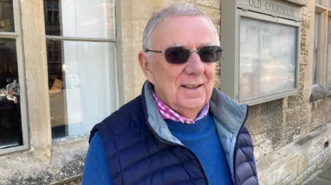 Glyn Williams. He has short grey hair and is wearing dark sunglasses, a blue jumper and a blue gilet. He is looking at the camera and smiling.
