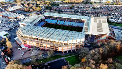 David Davies/PA Wire An aerial view of Aston Villa's Villa Park stadium. It is mostly white with a green pitch inside and light blue seating. Club banners on the front of the building are maroon in colour. Surrounding the stadium are car parks with cars, buildings and trees.