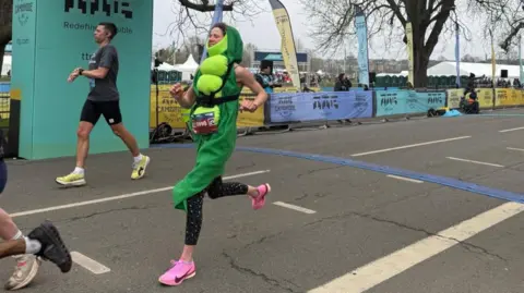 A woman dressed as a pea crossing the finishing line of the Cambridge Half Marathon. Beyond her is a man and beyond them both is the edge of a park with marquees and people milling around. 