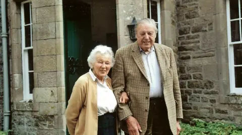 Palmer family Cherry Palmer, left, and Bill Palmer, right. Cherry is wearing a light brown cardigan, chequered skirt and white blouse, and Bill is wearing a patterned jacket, dark trousers and white shirt
