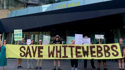 Protesters holding signs and a large yellow banner reading “Save Whitewebbs” demonstrate outside London’s City Hall against plans to build a Tottenham Hotspur training ground in the park.