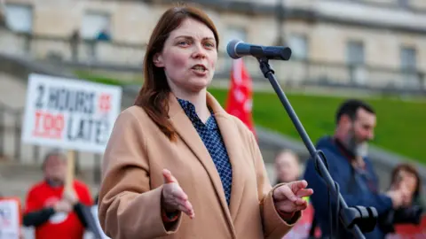 PA Media Sinn Féin MLA Jemma Dolan standing behind a microphone. She is wearing a blue top and a cream jacket. She has long brown hair. There are campaigners standing behind her. 